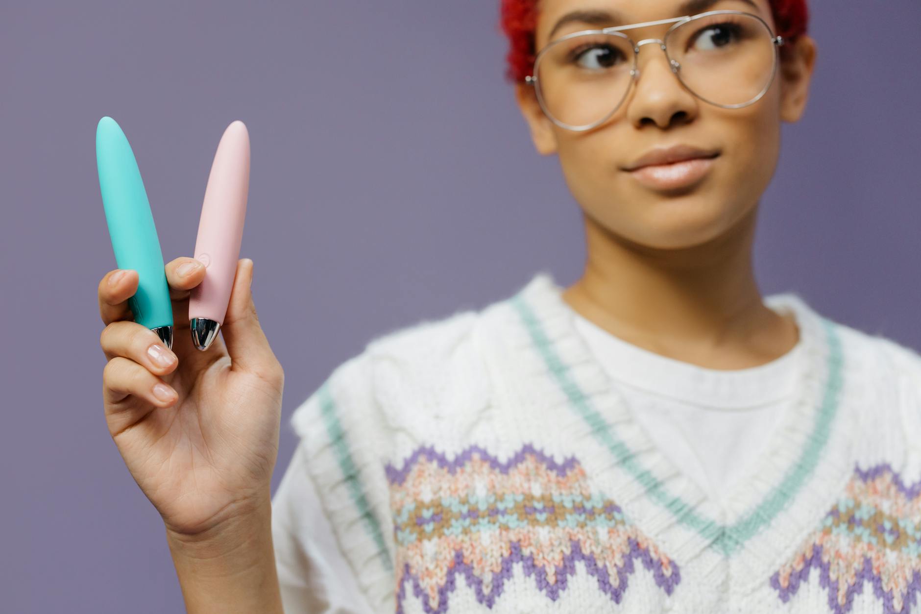 A young woman holding two vibrators against a purple background, representing different pleasure tools