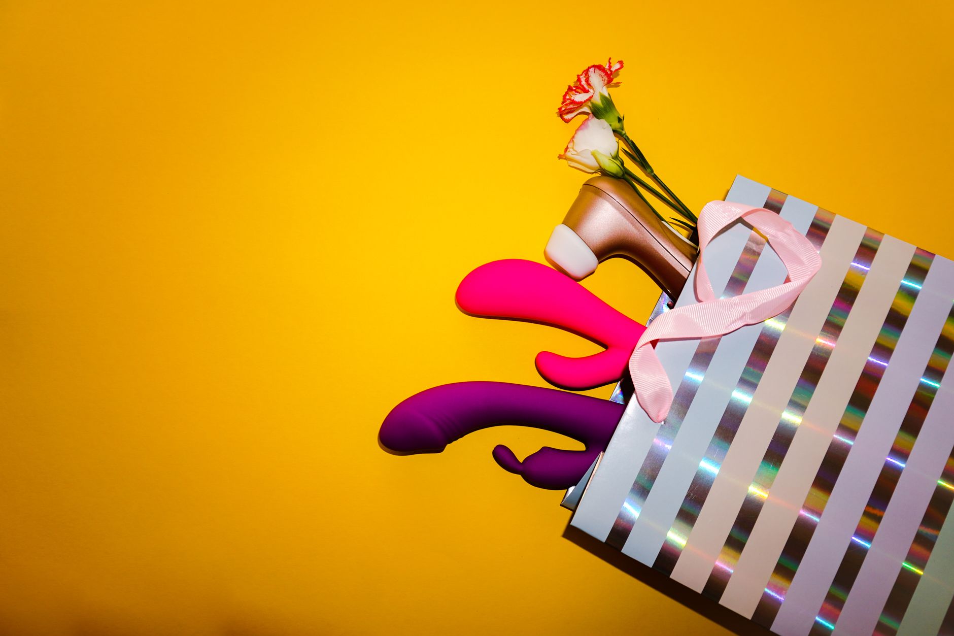 Colorful vibrators displayed in a holographic gift bag against a bold yellow background, representing accessible pleasure options.